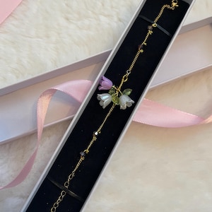 May include: A gold-coloured bracelet with miniature white and purple flower charms, green leaves, and tiny beads. The bracelet is displayed in a white gift box with a black interior, set against a fluffy white background.