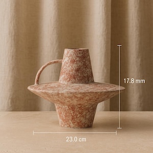 Puede incluir: Un jarrón de cerámica con un diseño único. El jarrón tiene una base ancha y plana y un asa. El exterior es de color marrón rojizo y crema moteado. El jarrón mide 23,0 cm de ancho y 1,8 cm de alto.