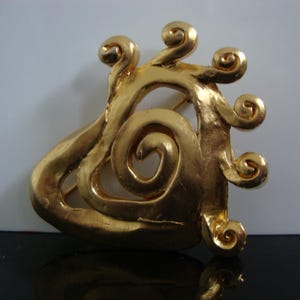 Op de afbeelding: Goudkleurige broche met een abstract ontwerp met wervelende, spiraalvormige vormen. De broche heeft een getextureerd oppervlak en een sluiting met speld. Het ontwerp doet denken aan de Art Nouveau-stijl.