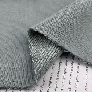 Pode incluir: Close-up de um tecido de malha cinza, mostrando a textura e a trama. O tecido está dobrado, revelando o verso com um padrão de malha diferente. As bordas são cruas e inacabadas. O tecido parece macio e flexível.