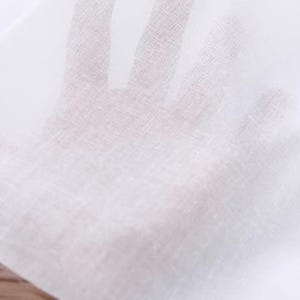 Puede incluir: Primer plano de una tela blanca transparente, posiblemente lino o algodón, sostenida a la luz. La tela es fina y translúcida, permitiendo que la mano detrás sea parcialmente visible. La textura parece ligeramente tejida.