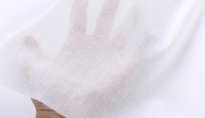 May include: Close-up of a sheer white fabric, possibly linen or cotton, held up to the light. The fabric is thin and translucent, allowing the hand behind it to be partially visible. The texture appears slightly woven.