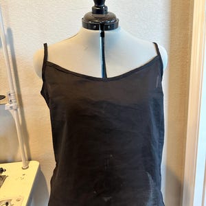 May include: A black camisole top with thin spaghetti straps and a scoop neckline. The top is displayed on a mannequin, showcasing its simple, elegant design. The fabric appears lightweight and slightly sheer.