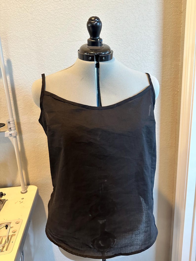 May include: A black camisole top with thin spaghetti straps and a scoop neckline. The top is displayed on a mannequin, showcasing its simple, elegant design. The fabric appears lightweight and slightly sheer.