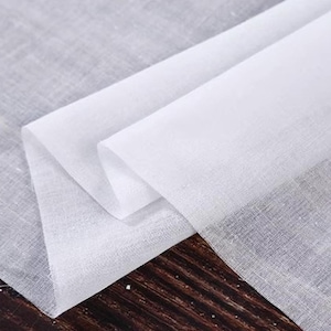 May include: Close-up of white linen fabric, folded and layered. The fabric has a slightly textured appearance and is semi-transparent. The folds create shadows, highlighting the fabric's texture. The fabric is likely used for clothing or home decor.