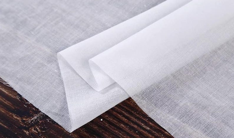 May include: Close-up of white linen fabric, folded and layered. The fabric has a slightly textured appearance and is semi-transparent. The folds create shadows, highlighting the fabric's texture. The fabric is likely used for clothing or home decor.