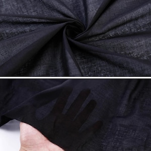 May include: Close-up of a dark navy blue sheer fabric. The fabric is lightweight and appears to be woven with a subtle texture. The fabric is draped and folded, creating shadows and depth. The fabric is held up to the light, revealing the hand behind it.