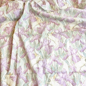 Puede incluir: Tela crema con un patrón repetido de lirios lavanda y conejos blancos. El diseño está realizado en estilo acuarela, creando una estética suave y vintage. Adecuado para diversos proyectos de manualidades.