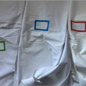 Puede incluir: Se muestran tres secciones de tela blanca, cada una con un peso escrito a mano en un rectángulo de color: 80g en verde, 150g en azul y 250g en rojo. La tela está doblada, mostrando variaciones de textura y peso.