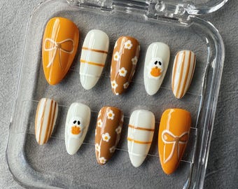 May include: A set of press-on nails in a clear plastic case. The nails feature various designs, including orange bows, white and orange stripes, brown daisy patterns, and a duck face. The nails are oval-shaped.