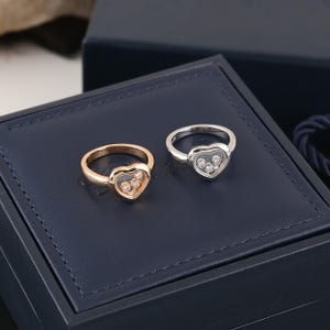 Könnte beinhalten: Zwei Ringe mit herzförmigen Designs. Einer ist goldfarben, der andere silberfarben. Jeder Ring hat ein herzförmiges Zentrum mit kleinen, klaren Steinen. Die Ringe werden auf einer dunkelblauen Oberfläche präsentiert.