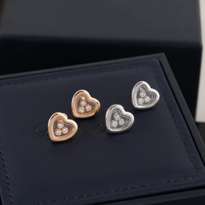 Puede incluir: Dos pares de pendientes de botón en forma de corazón. Un par es dorado, el otro plateado. Cada pendiente presenta tres pequeñas piedras transparentes suspendidas en el centro del corazón. Los pendientes se exhiben sobre una superficie azul oscuro.