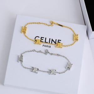Puede incluir: Dos pulseras, una dorada y otra plateada, se exhiben sobre una caja blanca. La pulsera dorada presenta una cadena con cuatro eslabones decorativos. La pulsera plateada tiene un diseño similar. La caja tiene el texto "CELINE PARIS".