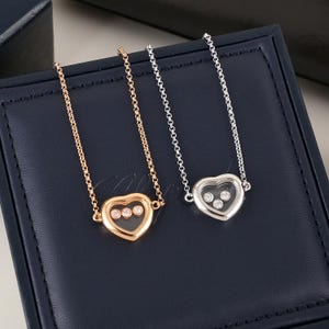 Könnte beinhalten: Zwei Halsketten mit herzförmigen Anhängern. Eine Halskette hat eine goldene Kette und einen Anhänger, die andere eine silberne Kette und einen Anhänger. Jeder Herzanhänger enthält drei kleine, klare Steine. Die Halsketten werden auf einer dunkelblauen Oberfläche präsentiert.