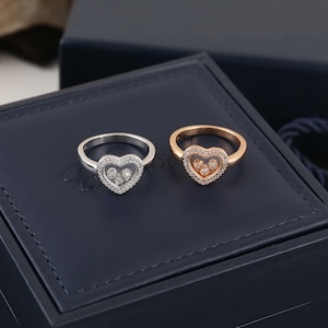 Könnte beinhalten: Zwei Ringe mit herzförmigen Designs. Ein Ring ist silberfarben mit einem klaren herzförmigen Zentrum, das mit kleinen, klaren Steinen gefüllt und von einer Reihe kleinerer Steine umgeben ist. Der andere Ring ist goldfarben mit einem ähnlichen Design.