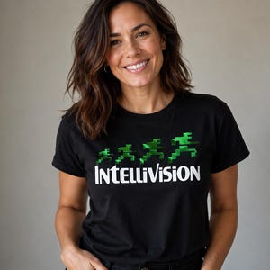 May include: A black short-sleeved t-shirt featuring the white text Intellivision printed across the chest. Above the text are four green pixelated figures in varying sizes, arranged in a sequence that suggests a running motion from left to right.