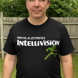 May include: Black t-shirt featuring the text "MATTEL ELECTRONICS INTELLIVISION" in white, with a green pixelated running figure on the right side. The shirt is worn by a person standing in front of a wooden fence.