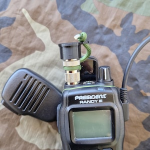 Puede incluir: Una radio CB President Randy III negra con una antena verde y negra y un cordón verde. La radio tiene una pantalla digital y un micrófono.