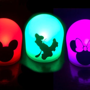 May include: Three small, round, battery-operated night lights with different colored lights and character silhouettes. The first light is red with a black Mickey Mouse silhouette. The second light is green with a black silhouette of a cartoon dog. The third light is purple with a black Minnie Mouse silhouette with a bow.