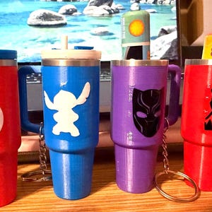 Könnte beinhalten: Vier bunte Reisebecher mit Griffen und Schlüsselanhängern. Die Becher sind rot, blau und lila und haben jeweils ein anderes Charakterdesign: das Avengers-Logo, Stitch und Black Panther. Auf einem befindet sich ein kleiner Behälter mit einem Traubenmuster.