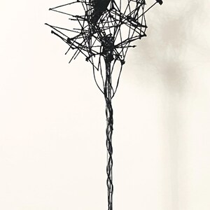 May include: A black wire sculpture of a tree with a twisted trunk and a complex, web-like canopy. The sculpture is mounted on a black square base with a pyramid-shaped top.