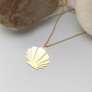 Könnte beinhalten: Eine goldfarbene Muschel-Anhänger-Halskette. Der Anhänger ist ein stilisiertes Muscheldesign mit mehreren gebogenen Linien. Die Halskette hat eine zarte, goldfarbene Kette. Die Halskette wird auf einer weißen Oberfläche mit einem hellen Stein im Hintergrund präsentiert.