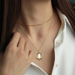May include: Gold-colored layered necklace. The top layer is a delicate chain, and the lower layer features a pendant with a tree silhouette inside a circle. The necklace is worn over a white button-down shirt.