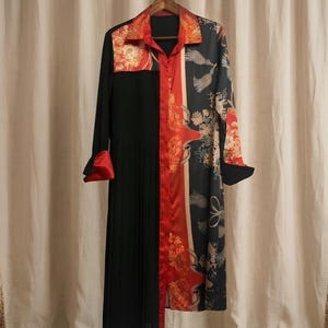 May include: A long-sleeved dress with a unique design. The dress features a black pleated skirt, a red and floral patterned panel, and a black and floral patterned sleeve. The collar and cuffs are red. The dress is hanging on a wooden hanger.