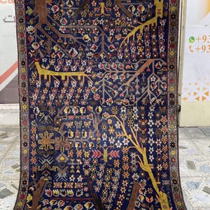 Könnte beinhalten: Ein rechteckiger, dunkelblauer Teppich mit einem detaillierten Lebensbaum-Design. Der Teppich hat braune Baumstämme, gelbe Äste und farbenfrohe Blumen- und geometrische Muster. Der Rand ist eine Mischung aus Farben.