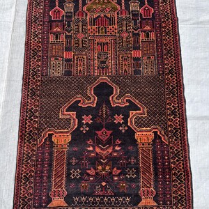May include: A rectangular, hand-woven rug with a detailed, symmetrical design. The rug features a dark background with intricate patterns in shades of red, orange, and brown. The design includes architectural elements and geometric shapes, with a fringed edge.