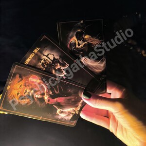 May include: A hand holds a fan of tarot cards with dark, detailed illustrations. The cards feature images of a lion, a couple embracing, and a chariot. The cards have gold borders and the word "Strength" is visible on one card. The background is dark.