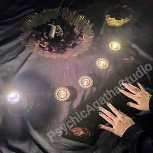 May include: A mystical scene featuring a deck of tarot cards with an illuminated symbol, surrounded by lit candles and a decorative bowl filled with petals. The text "PsychicAgathaStudio" is visible.