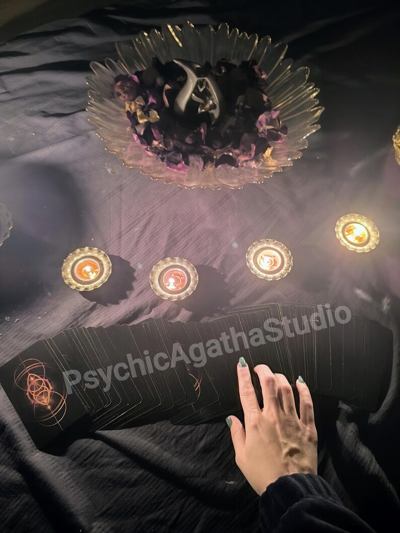 May include: A deck of black tarot cards with gold geometric designs, arranged on a dark surface. The text "PsychicAgathaStudio" is visible. A clear glass bowl with dried flowers and candles are also present.