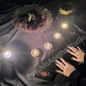 May include: A mystical scene featuring a deck of tarot cards, lit candles, and a decorative bowl with flower petals. The text "PsychicAgathaStudio" is visible. The scene is set on a dark, textured surface, creating a mysterious ambiance.