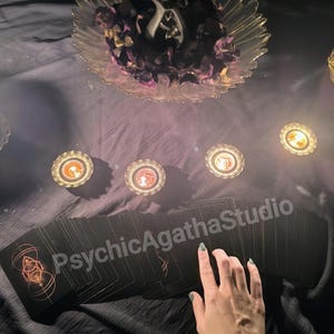 May include: A deck of black tarot cards with gold geometric designs, arranged on a dark surface. The text "PsychicAgathaStudio" is visible. A clear glass bowl with dried flowers and candles are also present.