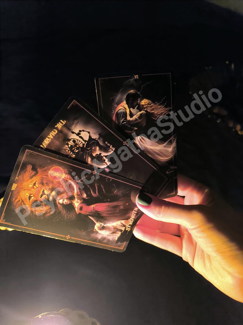 May include: A hand holds a fan of tarot cards with dark, detailed illustrations. The cards feature images of a lion, a couple embracing, and a chariot. The cards have gold borders and the word "Strength" is visible on one card. The background is dark.