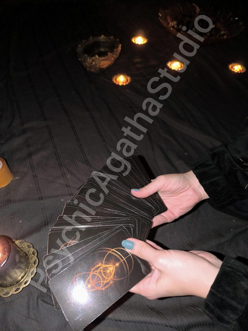 May include: A deck of black tarot cards with a gold geometric design is held by hands with blue nail polish. The cards are fanned out against a dark background with lit candles and decorative objects. The text "Psychic Agatha Studio" is visible.