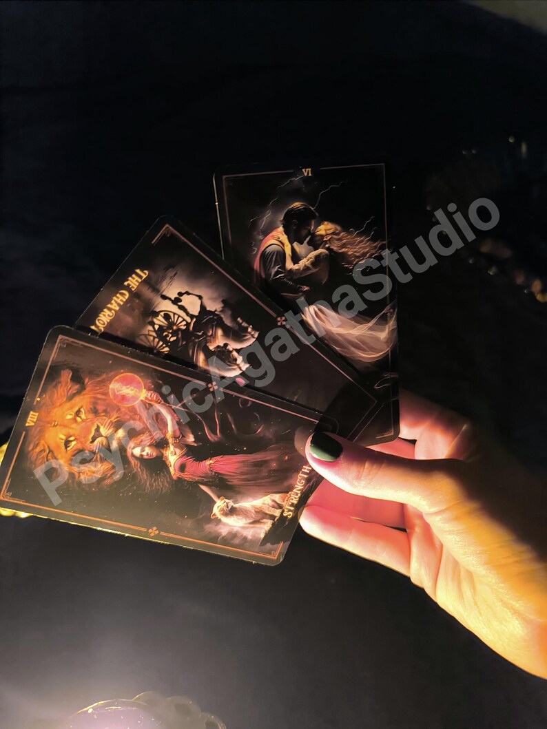 May include: A hand holds a fan of tarot cards with dark, detailed illustrations. The cards feature images of a lion, a couple embracing, and a chariot. The cards have gold borders and the word "Strength" is visible on one card. The background is dark.