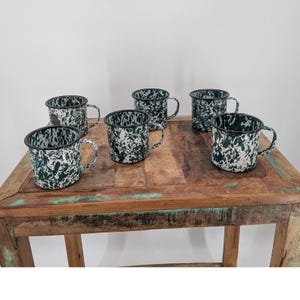 May include: A set of five mugs featuring a white and dark green speckled enamel finish. Each mug has a cylindrical body, a side handle, and a dark rim. The mugs are arranged in a group, showcasing the consistent mottled pattern across all pieces.