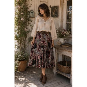 May include: A long-sleeved beige blouse with a gathered neckline and lace trim on the cuffs, paired with a dark, calf-length skirt featuring a floral pattern in shades of burgundy, black, and white, cinched with a wide brown leather belt.