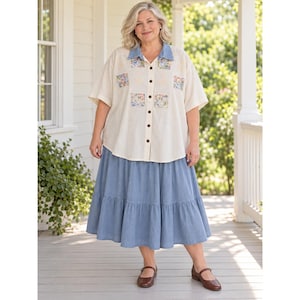May include: A cream-coloured button-down shirt with short sleeves, a denim-style collar, and four square floral patches on the front. The shirt is paired with a calf-length, tiered blue skirt and brown leather Mary Jane shoes.