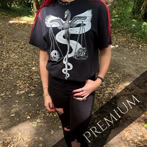 Op de afbeelding: Een zwart t-shirt met korte mouwen en een witte grafische print van een zwaan met gespreide vleugels die een weegschaal vasthoudt. Aan de ene kant van de weegschaal ligt een menselijke schedel, aan de andere kant een anatomisch hart, verbonden door een kronkelende slang.