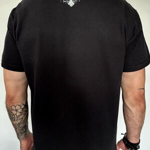 Op de afbeelding: Een zwart t-shirt met korte mouwen en een witte afbeelding op de bovenrug. De afbeelding toont een gestileerde vogel met gespreide vleugels en een schedelachtige kop, vergezeld van de tekst "The Last Savior" in een gotisch lettertype.