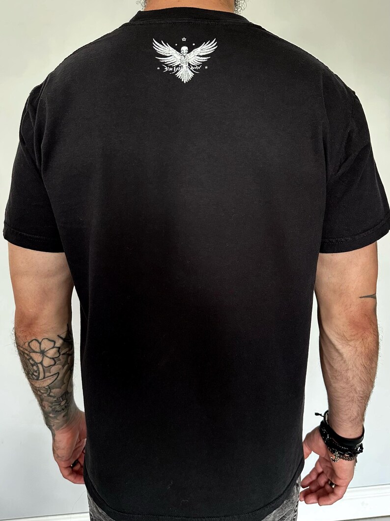 Op de afbeelding: Een zwart t-shirt met korte mouwen en een witte afbeelding op de bovenrug. De afbeelding toont een gestileerde vogel met gespreide vleugels en een schedelachtige kop, vergezeld van de tekst "The Last Savior" in een gotisch lettertype.