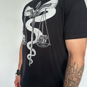 Op de afbeelding: Een zwart T-shirt met korte mouwen en een witte grafische print van een gevleugeld zwaard verstrengeld met een slang, die een weegschaal vormt. Aan de ene kant van de weegschaal ligt een menselijke schedel, aan de andere kant een menselijk hart.