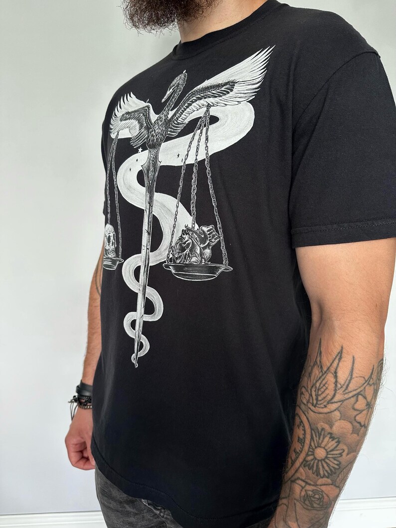 Op de afbeelding: Een zwart T-shirt met korte mouwen en een witte grafische print van een gevleugeld zwaard verstrengeld met een slang, die een weegschaal vormt. Aan de ene kant van de weegschaal ligt een menselijke schedel, aan de andere kant een menselijk hart.