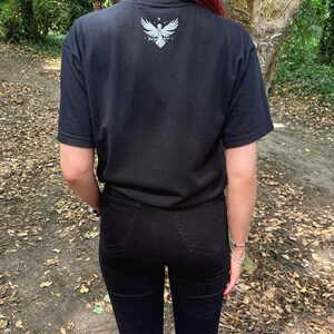 Op de afbeelding: Een zwart T-shirt met korte mouwen en een witte afbeelding van een gevleugeld vogelembleem in het midden op de bovenrug. Het shirt wordt gecombineerd met een zwarte spijkerbroek. De afbeelding bevat een klein sterretje boven de vogel en tekst.