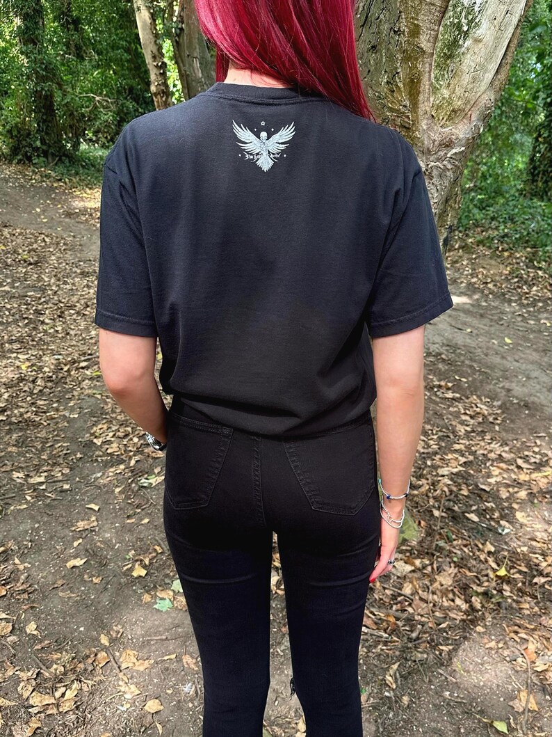 Op de afbeelding: Een zwart T-shirt met korte mouwen en een witte afbeelding van een gevleugeld vogelembleem in het midden op de bovenrug. Het shirt wordt gecombineerd met een zwarte spijkerbroek. De afbeelding bevat een klein sterretje boven de vogel en tekst.