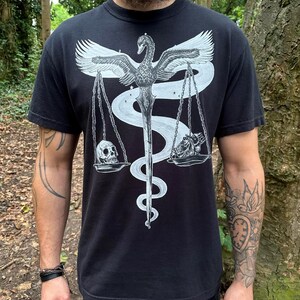 Op de afbeelding: Een zwart t-shirt met korte mouwen en een witte grafische print van een zwaan met gespreide vleugels, boven een zwaard dat omstrengeld is door een slang. Het zwaard ondersteunt een weegschaal met een menselijke schedel aan de ene kant en een hart aan de andere kant.