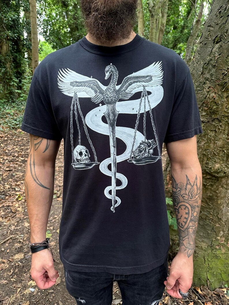 Op de afbeelding: Een zwart t-shirt met korte mouwen en een witte grafische print van een zwaan met gespreide vleugels, boven een zwaard dat omstrengeld is door een slang. Het zwaard ondersteunt een weegschaal met een menselijke schedel aan de ene kant en een hart aan de andere kant.